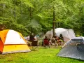 Grupa przyjaciół odpoczywa na polu namiotowym Mazury. Wokół rozstawione są namioty, a na stole piknikowym stoją napoje i przekąski.