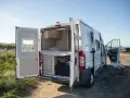 Biały kamper z otwartymi drzwiami, ukazujący łóżko i schowki. Idealne miejsce na życie w kamperze nad morzem.
