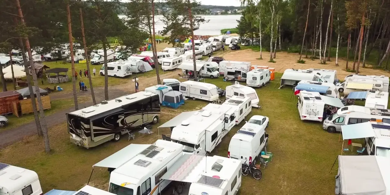 Duży zlot caravaningowy nad jeziorem. Wiele kamperów i przyczep kempingowych zaparkowanych na trawie, otoczonych drzewami.