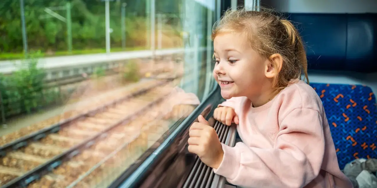 Uśmiechnięta dziewczynka patrzy przez okno pociągu. Zastanawia się, od ilu lat można jeździć samemu pociągiem, podziwiając mijane krajobrazy.