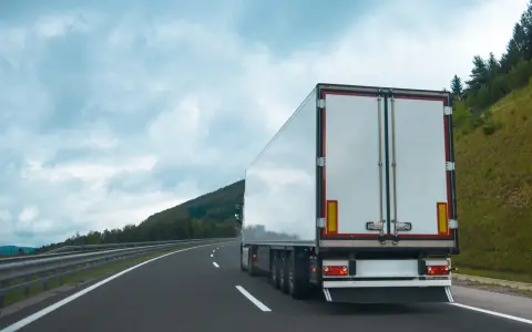 Ciężarówka na autostradzie, z tyłu widoczne białe drzwi naczepy. W tle góry i chmury. Tablice wyróżniające przepisy są częścią oznakowania pojazdu.