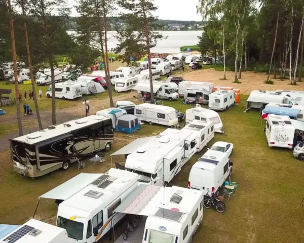 Duży zlot caravaningowy nad jeziorem. Wiele kamperów i przyczep kempingowych zaparkowanych na trawie, otoczonych drzewami.