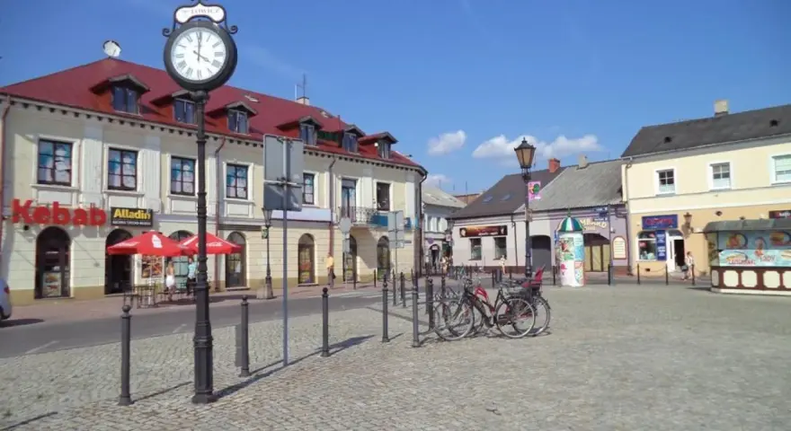 Słoneczny stary rynek łowicz z brukowanym placem, budynkami z czerwonymi dachami i zegarem na słupie.