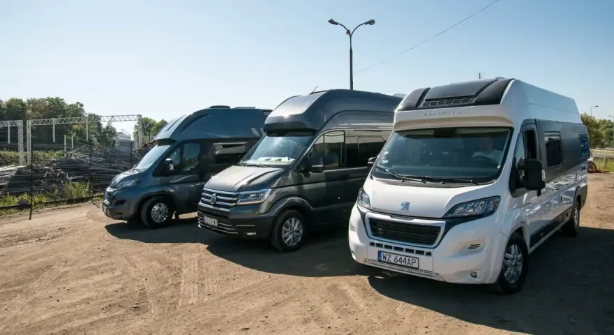 Trzy kampery stoją obok siebie na placu. Jeden z nich to kamper VW w ciemnoszarym kolorze.