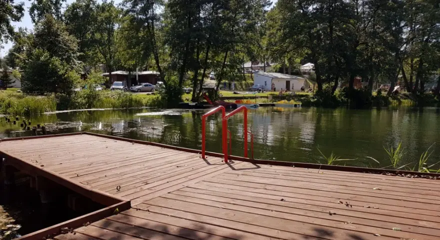 Drewniany pomost z czerwonymi poręczami nad jeziorem. W tle drzewa, budynki i samochody. Idealne miejsce na łagów camping.