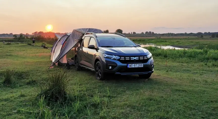 Dacia Jogger Camper rozłożony na biwaku o zachodzie słońca. Namiot połączony z autem, otoczony łąką i wodą.