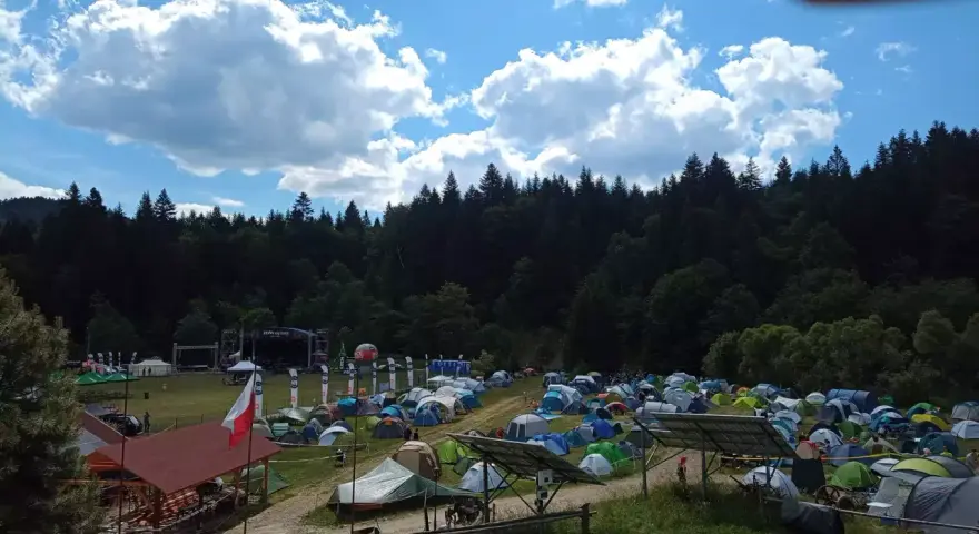 Pole namiotowe na festiwalu w Solinie. Tysiące namiotów pod błękitnym niebem z chmurami, w tle las i scena festiwalowa.