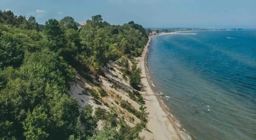 Mechelinki klif porośnięty drzewami, z piaszczystą plażą i turkusowym morzem. W oddali widać zabudowania i falochron.