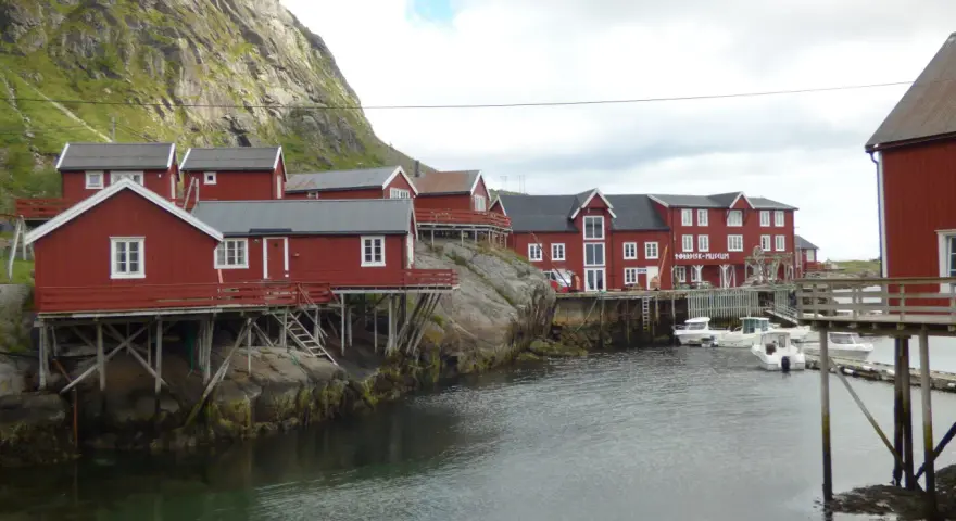 Czerwone domki rybackie na palach, przycumowane łodzie i skaliste zbocze tworzą malowniczy krajobraz Moskenes. Idealne miejsce na camping.