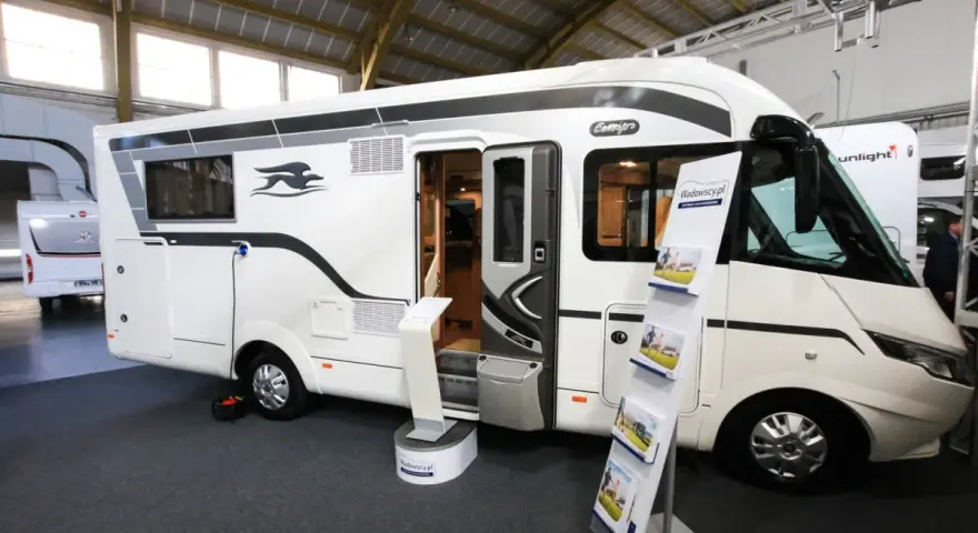 Biała kamperka z otwartymi drzwiami na targach caravaningowych Poznań. W środku widać wyposażenie, a na zewnątrz stojaki z ulotkami.