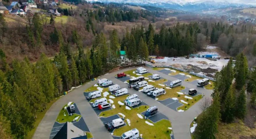 Widok z lotu ptaka na parking kempingowy ustup z wieloma kamperami i przyczepami. W tle góry i las.
