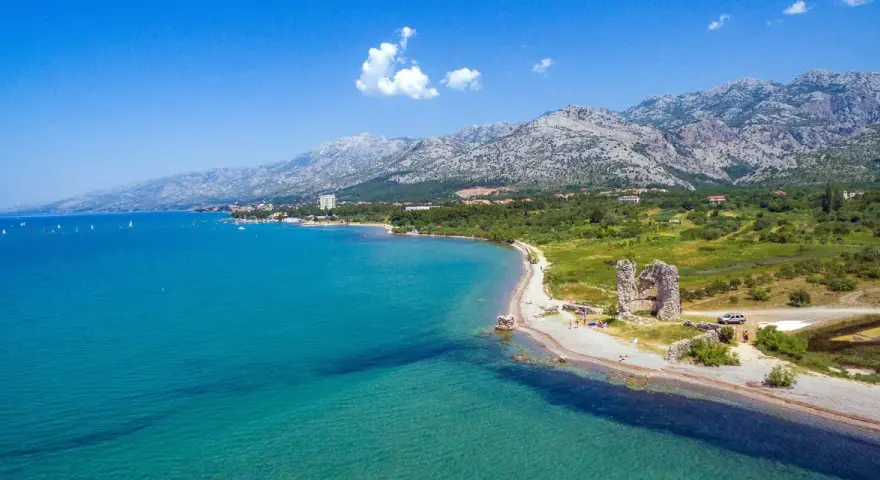 Piękne wybrzeże z górami w tle, idealne na camping Paklenica. Woda kusi turkusowym kolorem, a na plaży widać ruiny starej twierdzy.