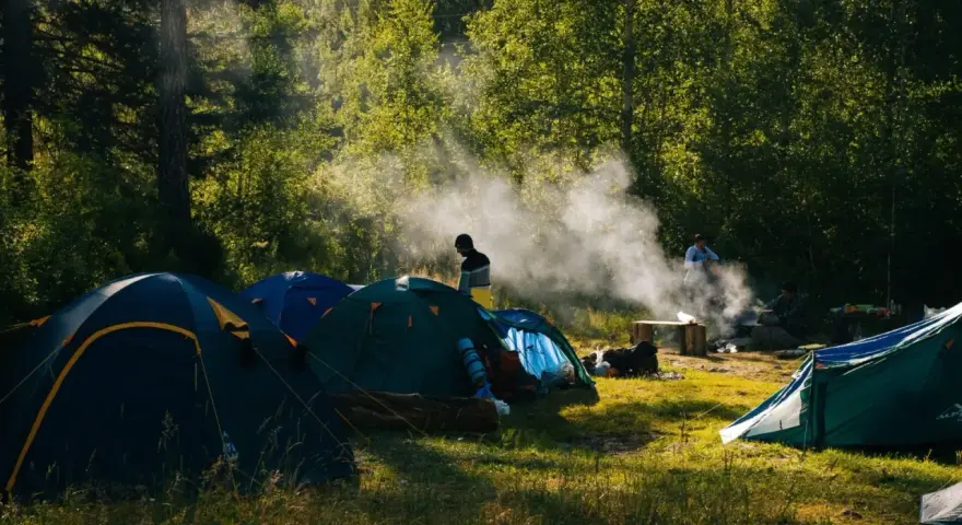 Słoneczny poranek na polu biwakowym. Kilka namiotów rozbitych wśród drzew, unosi się dym z ogniska.