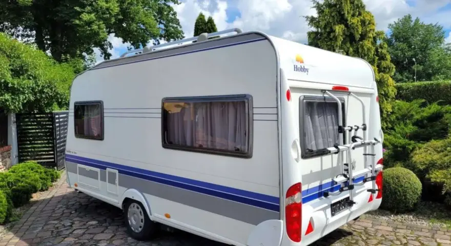 Biała przyczepa kempingowa Hobby 440 z niebieskimi pasami, zaparkowana na brukowanym podjeździe. Idealna do podróżniczego hobby.