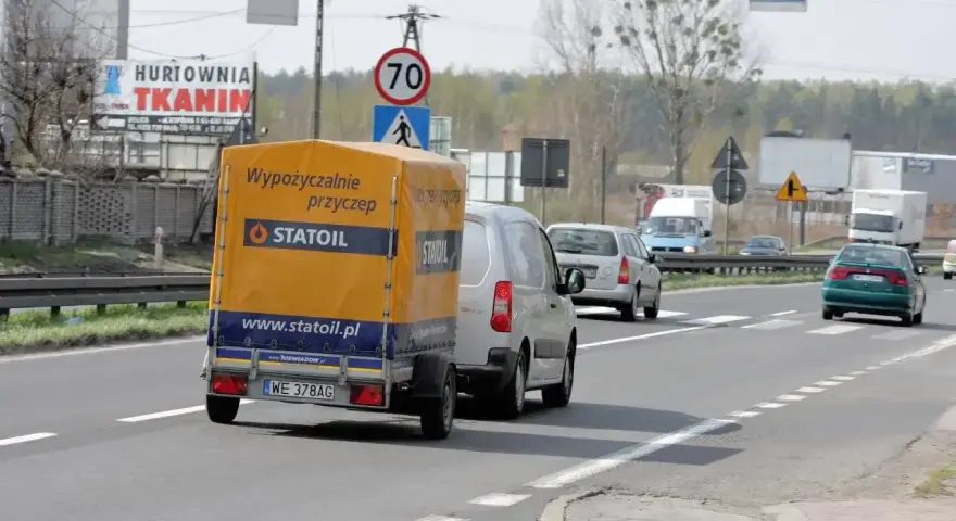 Samochód z żółtą przyczepką Statoil jedzie drogą. Znak ograniczenia do 70 km/h sugeruje, ile można jechać z przyczepką w tym miejscu.