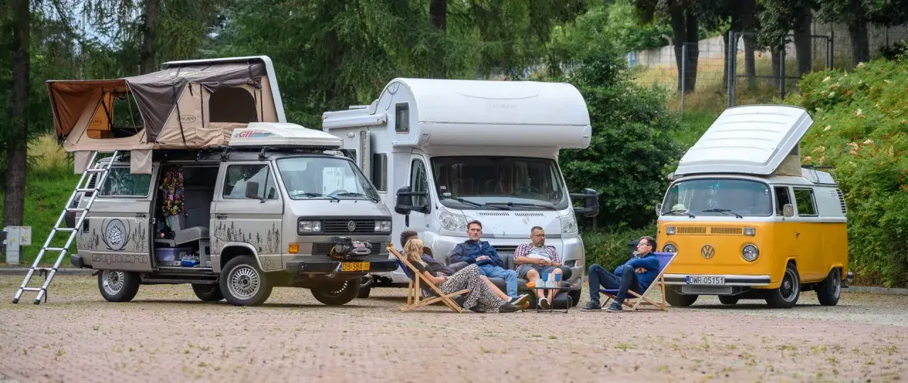 Grupa przyjaciół odpoczywa na leżakach przed trzema kamperami. Klasyczny żółty bus, srebrny van z namiotem dachowym i biała kamperka tworzą malowniczy parking dla kamperów.