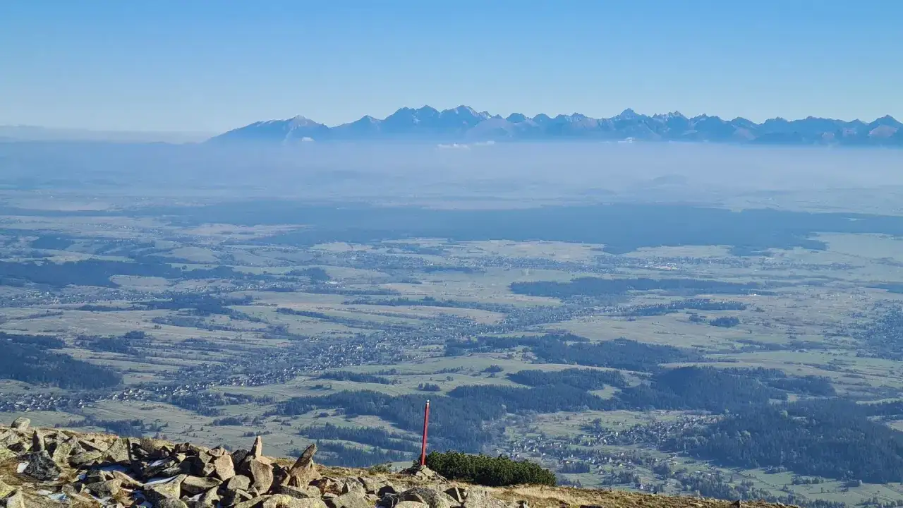 Widok z Babiej Góry na Tatry. Mgła otula doliny, a szczyty górskie rysują się na tle błękitnego nieba.