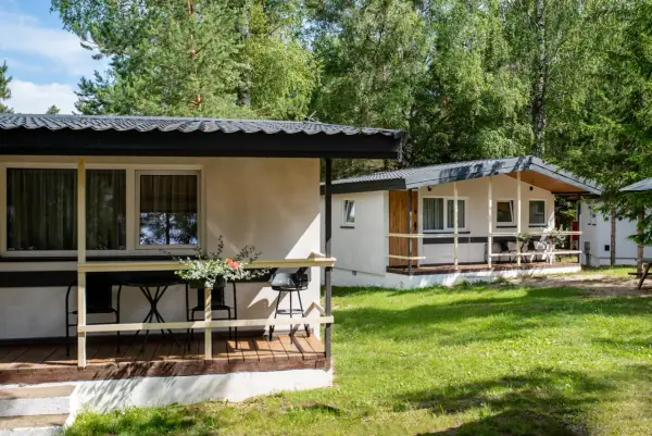 Dwa białe domki kempingowe w lesie, z werandami i meblami ogrodowymi. Idealne miejsce dla każdego **camping wagabunda**.