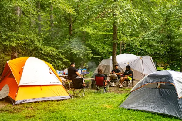 Grupa przyjaciół odpoczywa na polu namiotowym Mazury. Wokół rozstawione są namioty, a na stole piknikowym stoją napoje i przekąski.