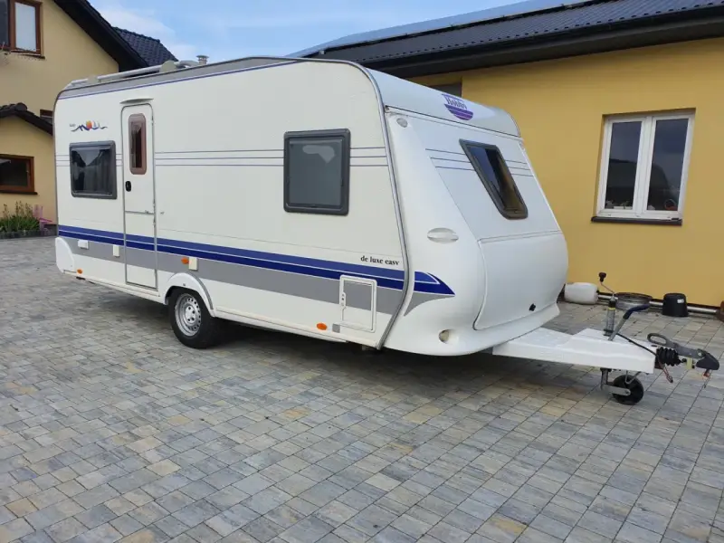 Biała przyczepa kempingowa Hobby 460 UFE "de luxe easy" stoi na kostce brukowej przed żółtym budynkiem.
